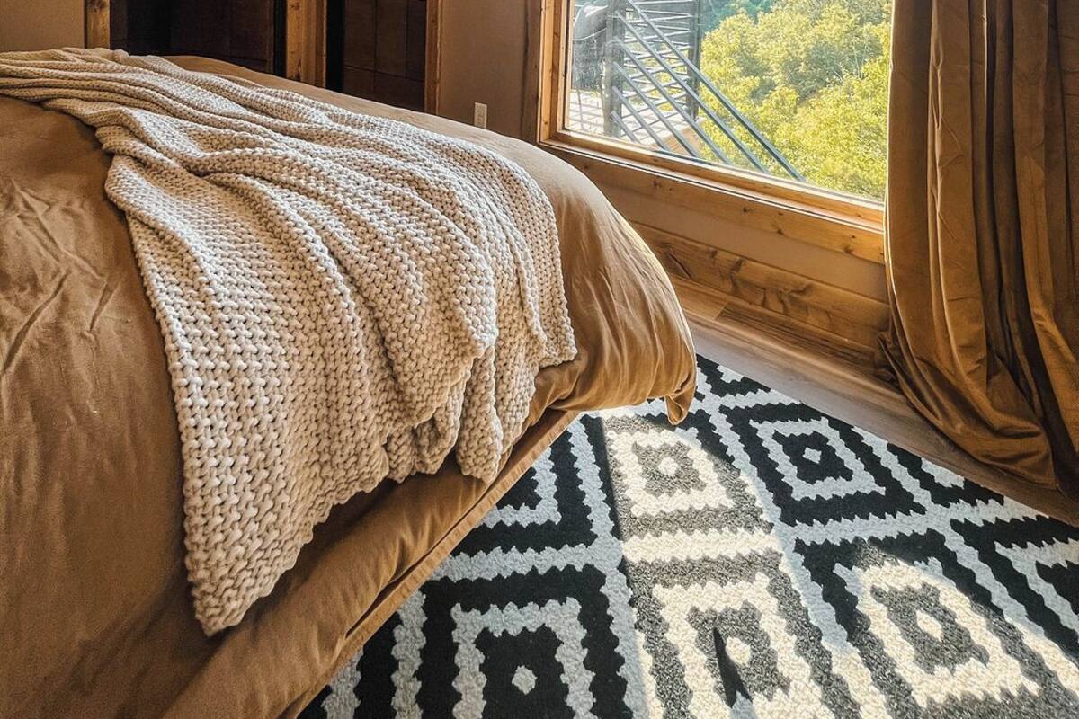 Un dormitorio con una cama grande, vistas a un bosque por una ventana y una alfombra estampada.