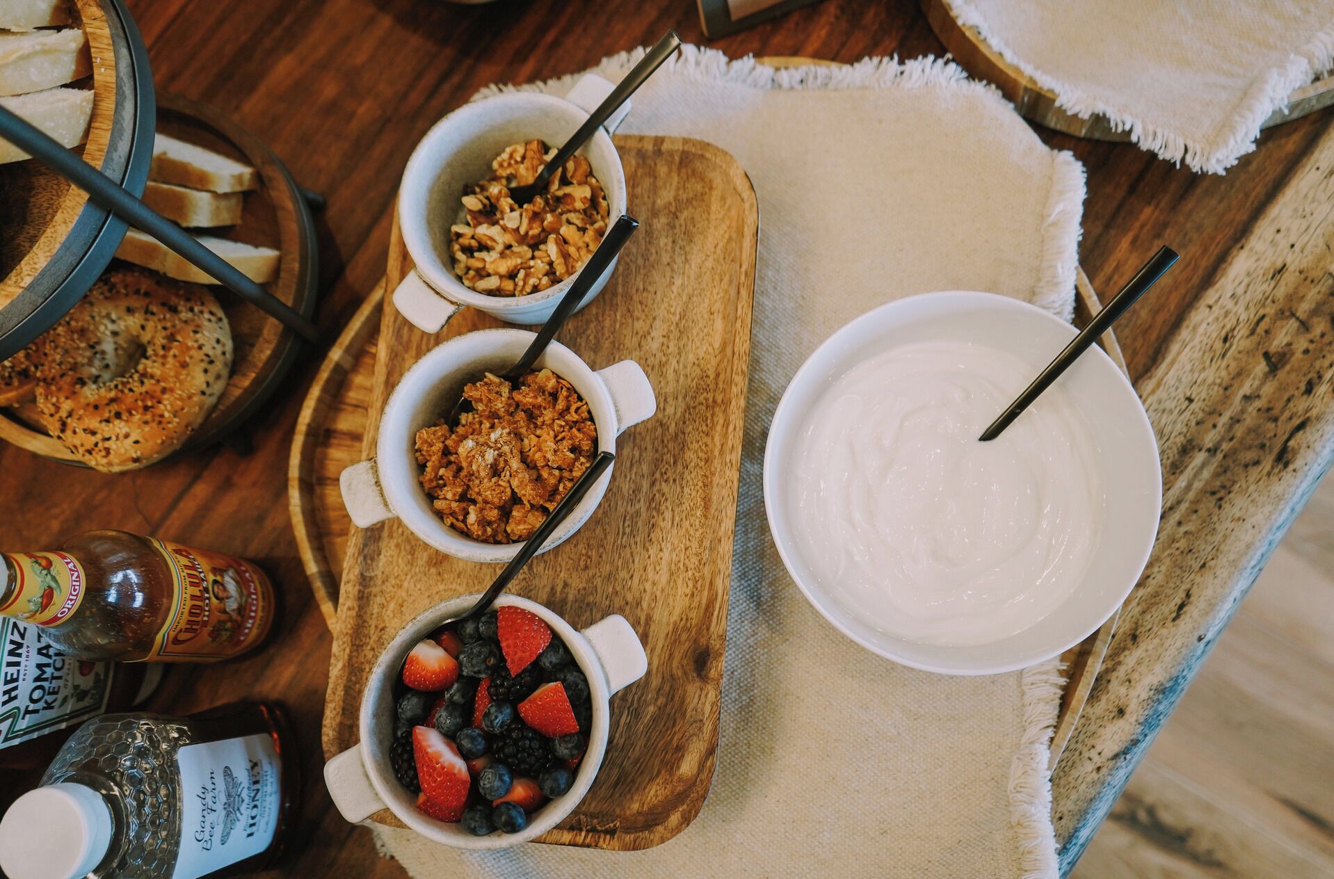 Une table dressée pour le petit-déjeuner, avec des bols de granola, de yaourt et des fruits, ainsi que des bouteilles de boissons sur un plateau en bois.