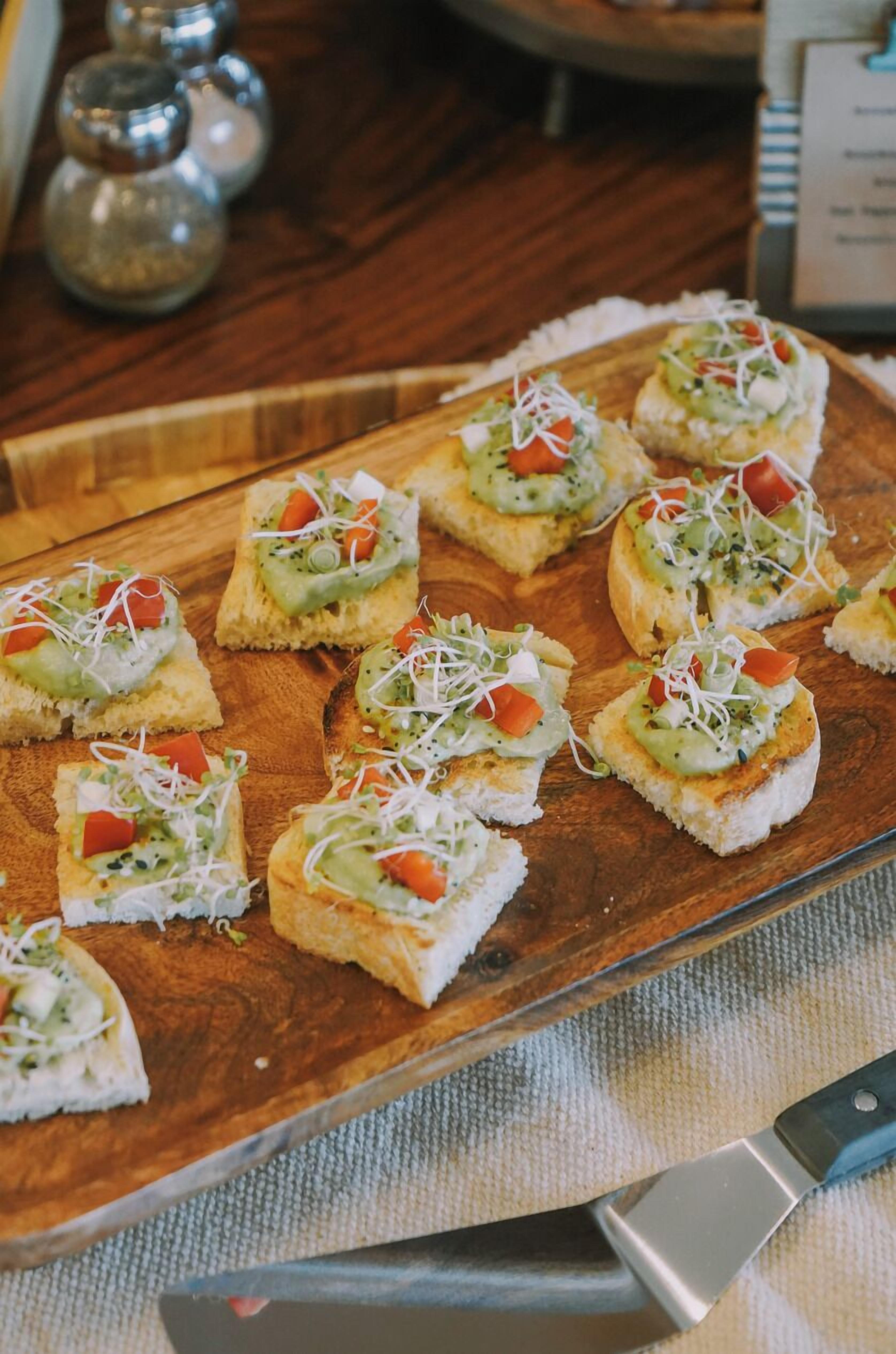 A wooden board with several pieces of toast topped with avocado, tomato, and sprouts.