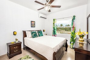 A bedroom with a bed, a nightstand, a dresser, a ceiling fan, an air conditioning unit, a window with curtains, and a view of the outdoors.