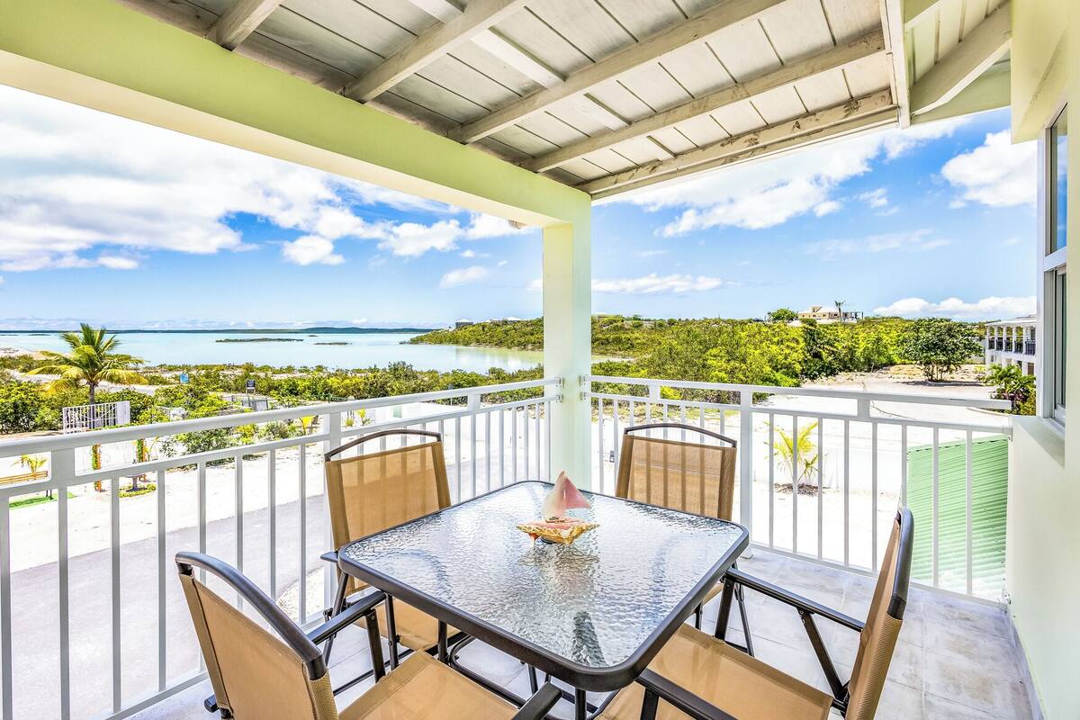 Un balcon extérieur abrité, doté d’une table en verre et de quatre chaises, offrant une vue sur une plage et l’océan.