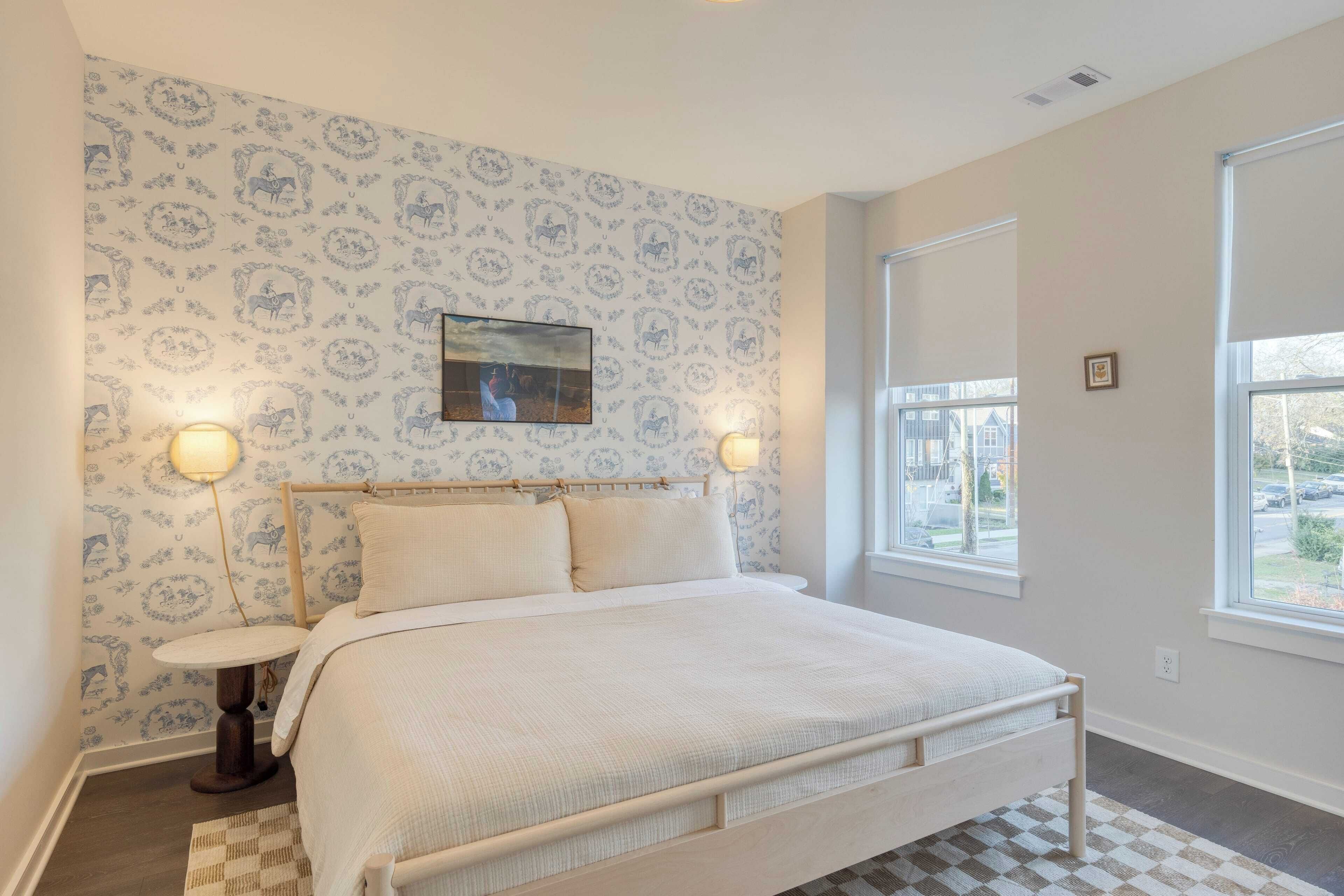 A bedroom with a bed, two bedside lamps, a patterned wallpaper, a framed picture on the wall, and a window with a view.