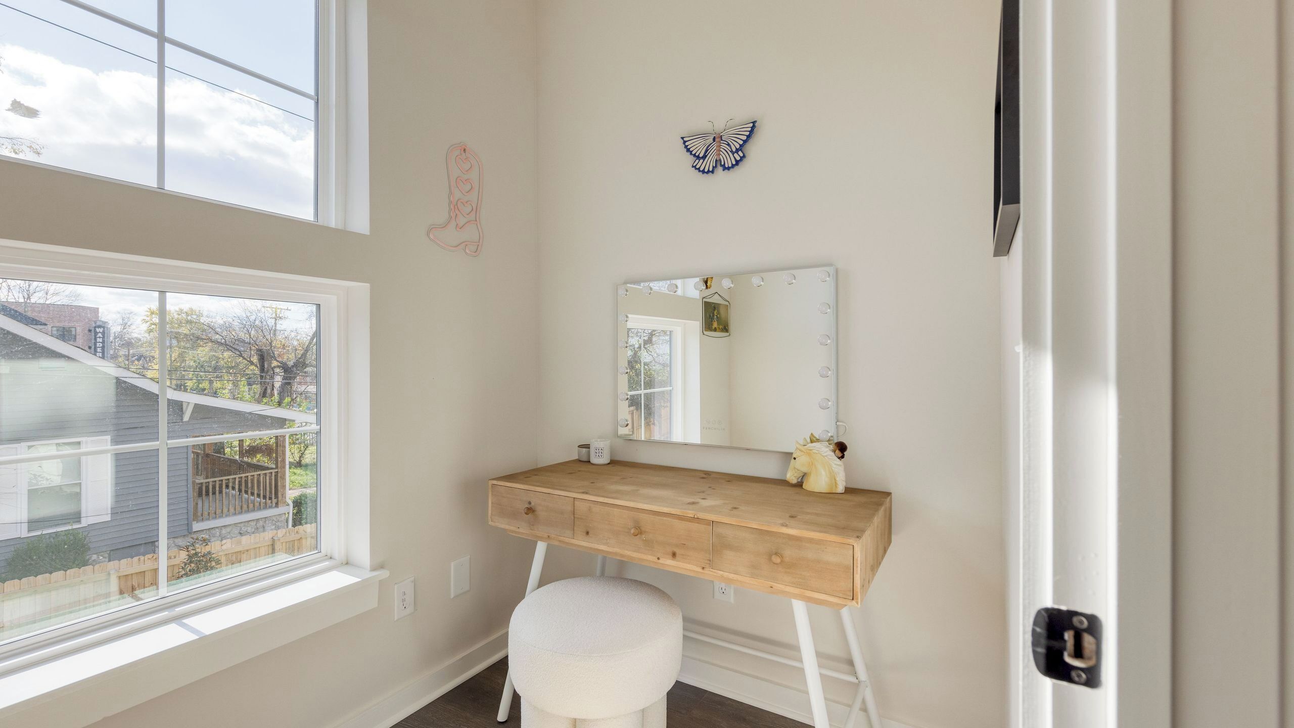 A room with a wooden vanity, a round stool, a large mirror with lights, and a window with a view of a house and trees.