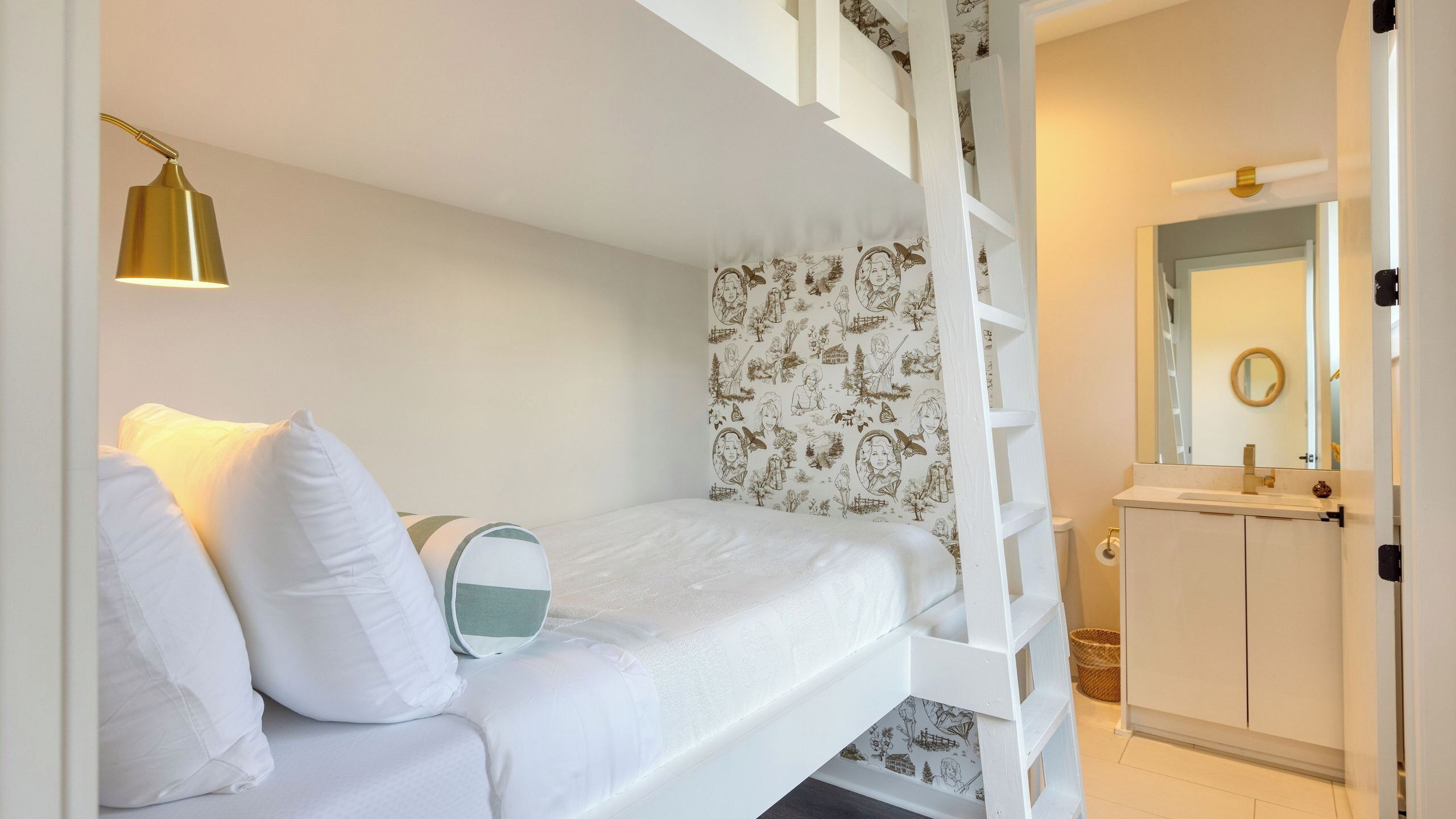 A bedroom with a bunk bed, a bedside lamp, a patterned wallpaper, a white wardrobe, and a bathroom with a mirror and sink.