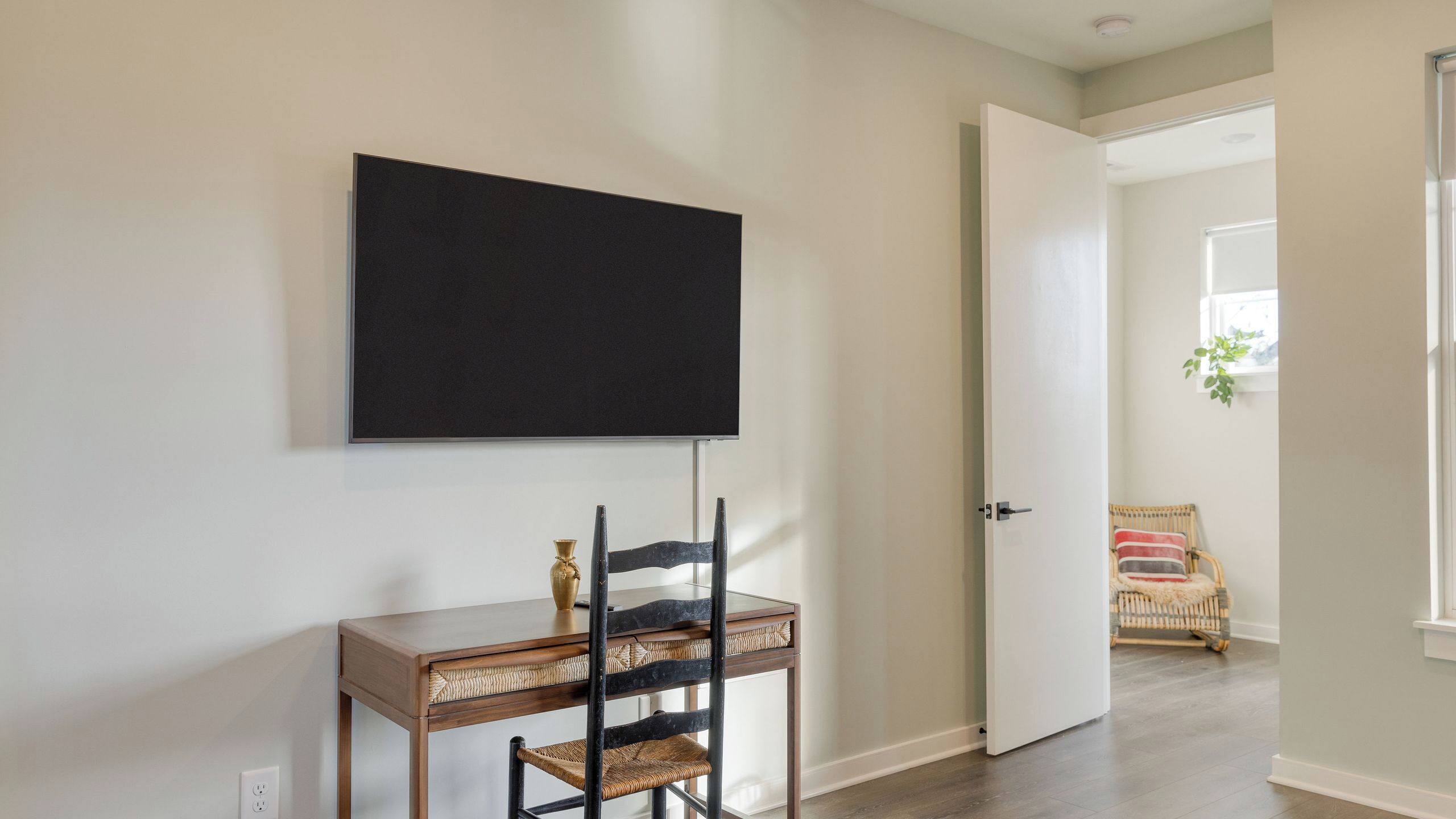 A room with a flat-screen TV mounted on the wall, a wooden table with a woven chair, and a door leading to another room.