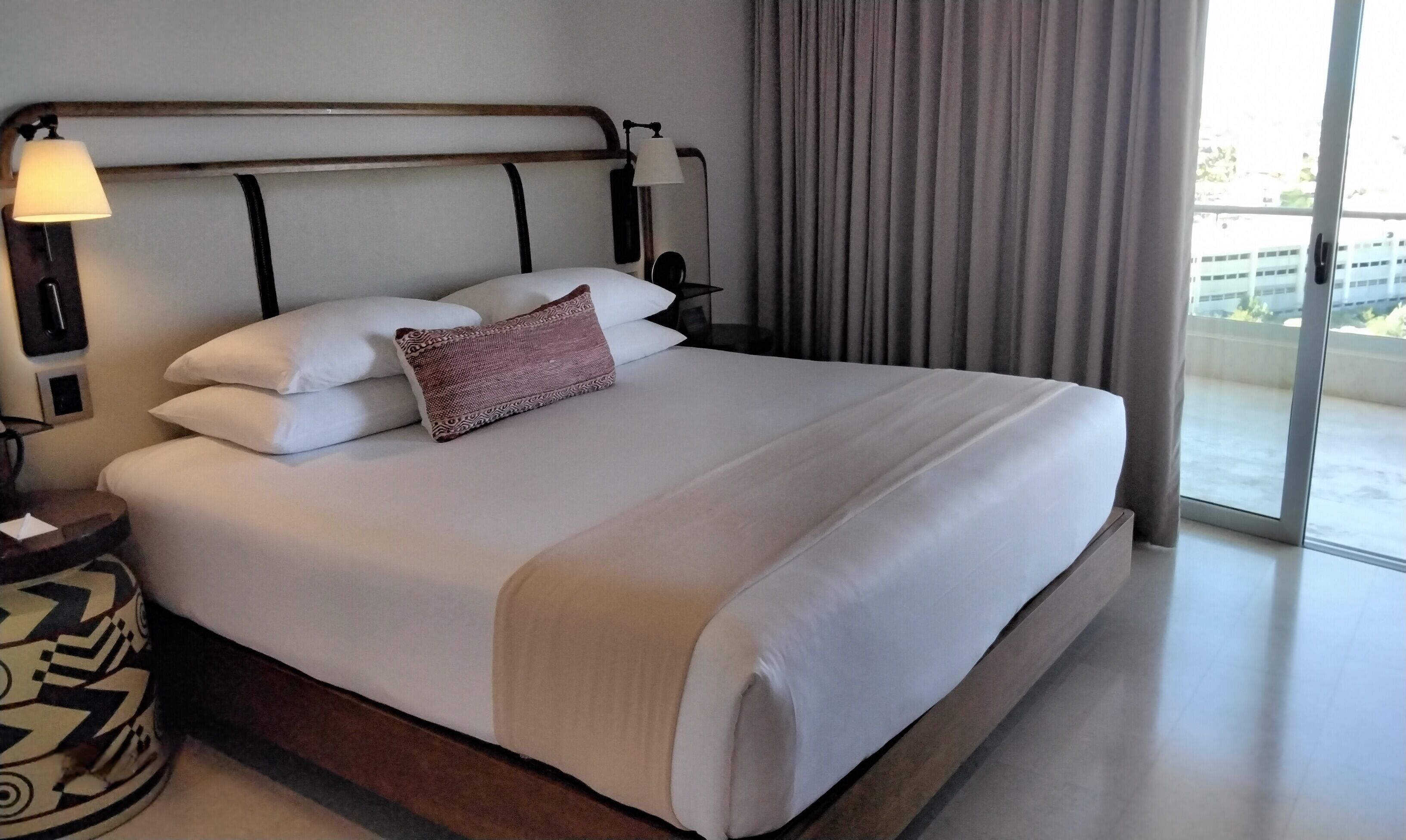 A hotel room with a large bed, a bedside lamp, a nightstand, and a view of the outside.