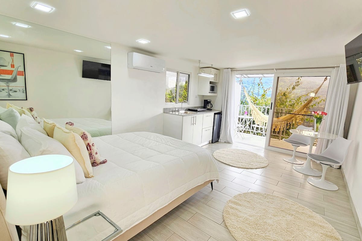 A modern bedroom with a large bed, a bedside table with a lamp, a small kitchen area, and a balcony with a hammock.