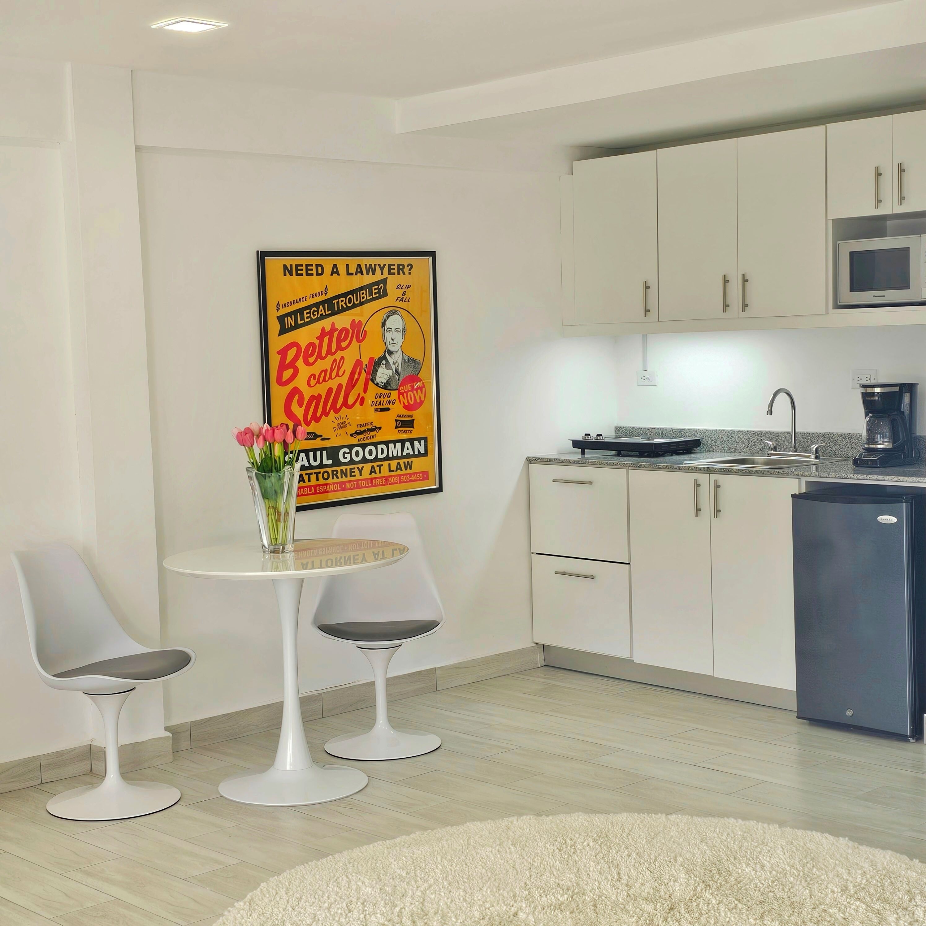 A modern kitchen with white cabinets, a microwave, and a coffee maker. There is a small dining area with two chairs and a table. A poster is visible on the wall.