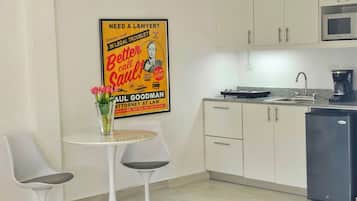 A modern kitchen with white cabinets, a microwave, and a coffee maker. There is a small dining area with two chairs and a table. A poster is visible on the wall.