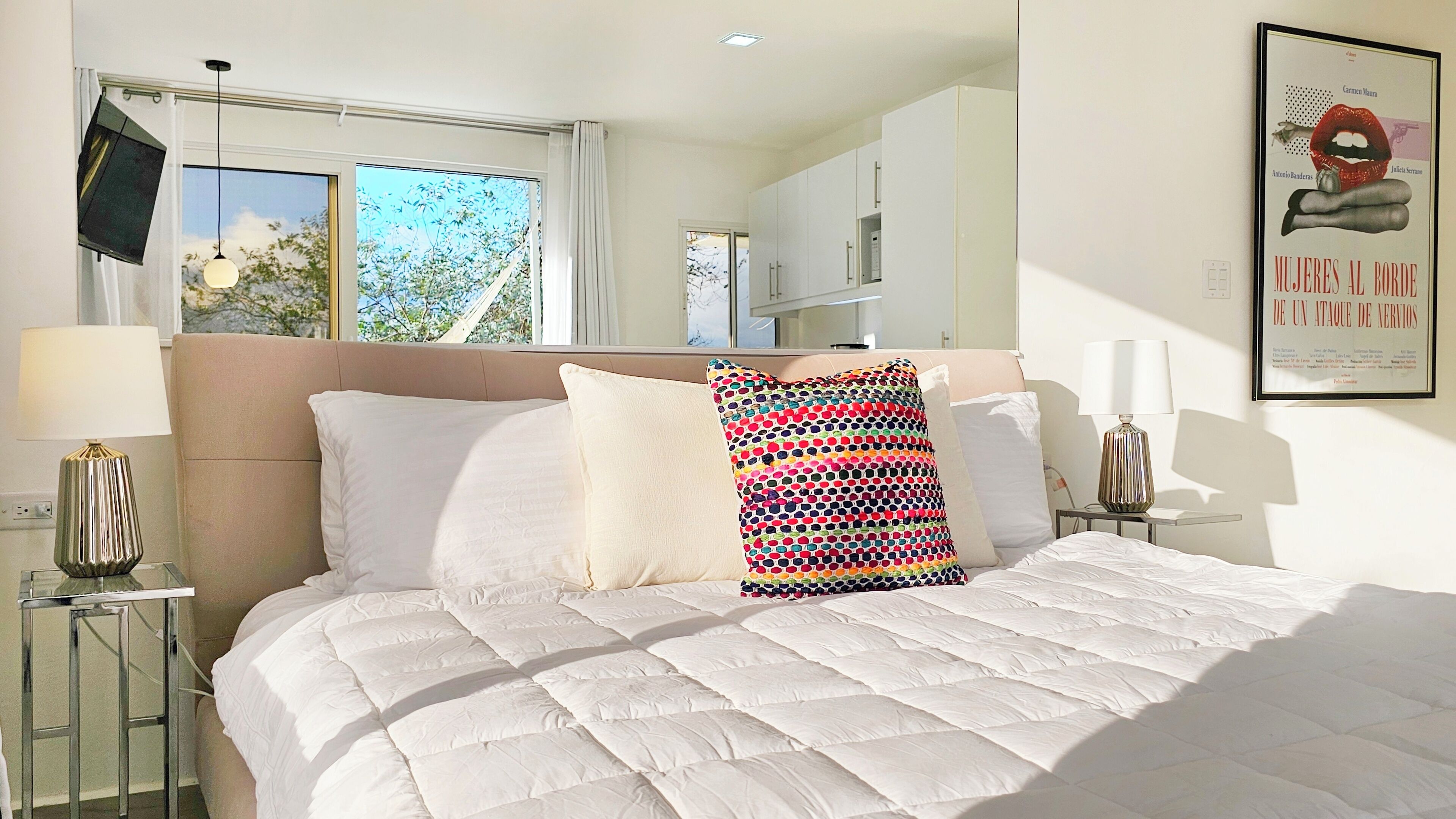 A bedroom with a large bed, a window with a view of trees, a kitchen area, and a framed artwork on the wall.