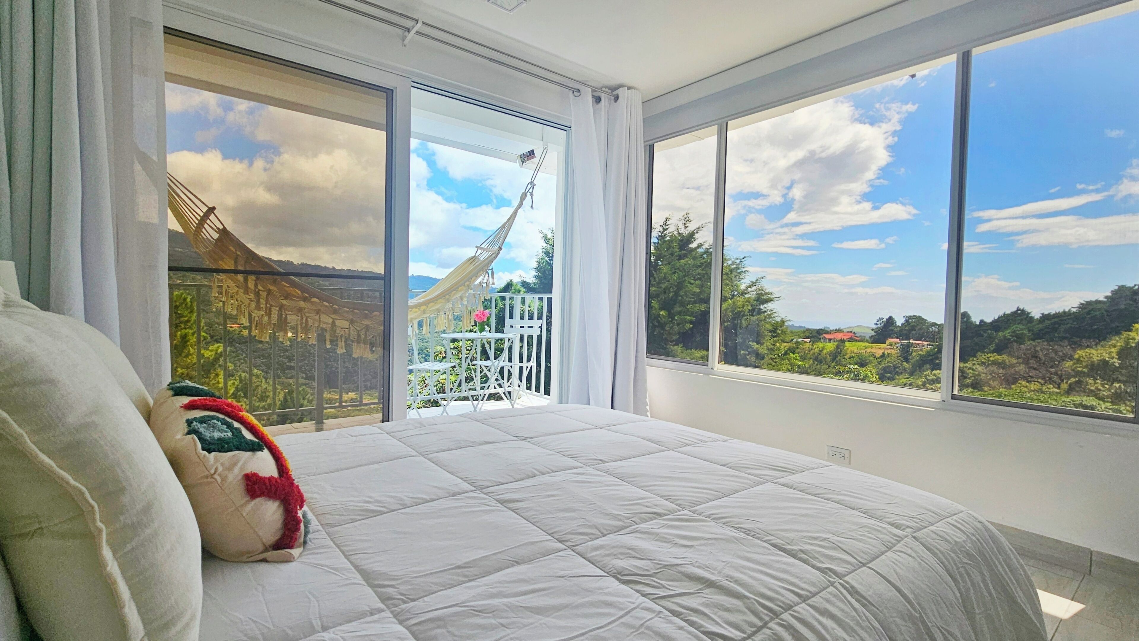Un dormitorio con una cama grande, vistas a un puente y un balcón con una hamaca y flores.