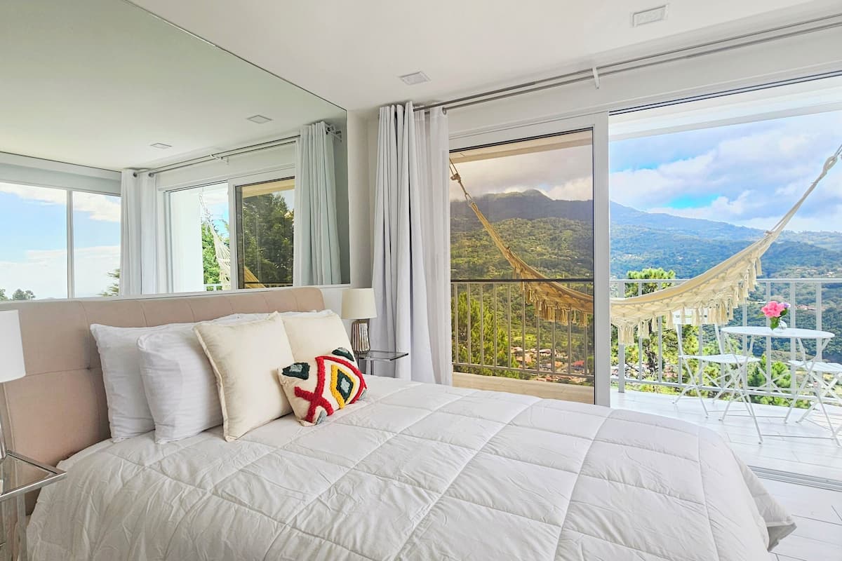 A spacious bedroom with a large bed, a nightstand, and a view of a balcony with a hammock and outdoor seating.