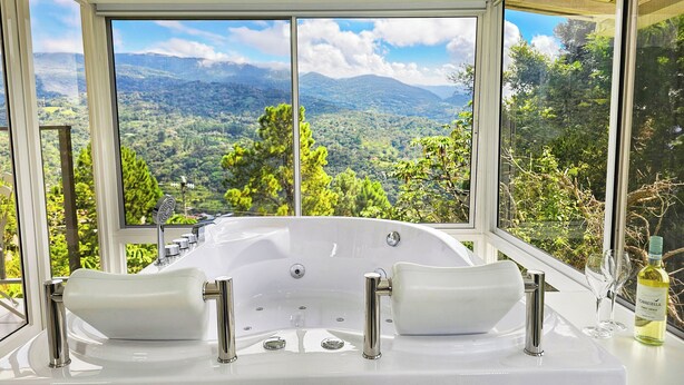 Eine große, weiße freistehende Badewanne mit mehreren Wasserhähnen und Blick auf eine gebirgige Landschaft durch große Fenster.