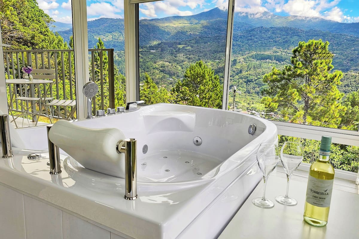 A bathroom with a large bathtub, a window with a mountain view, and a bottle of wine on the counter.