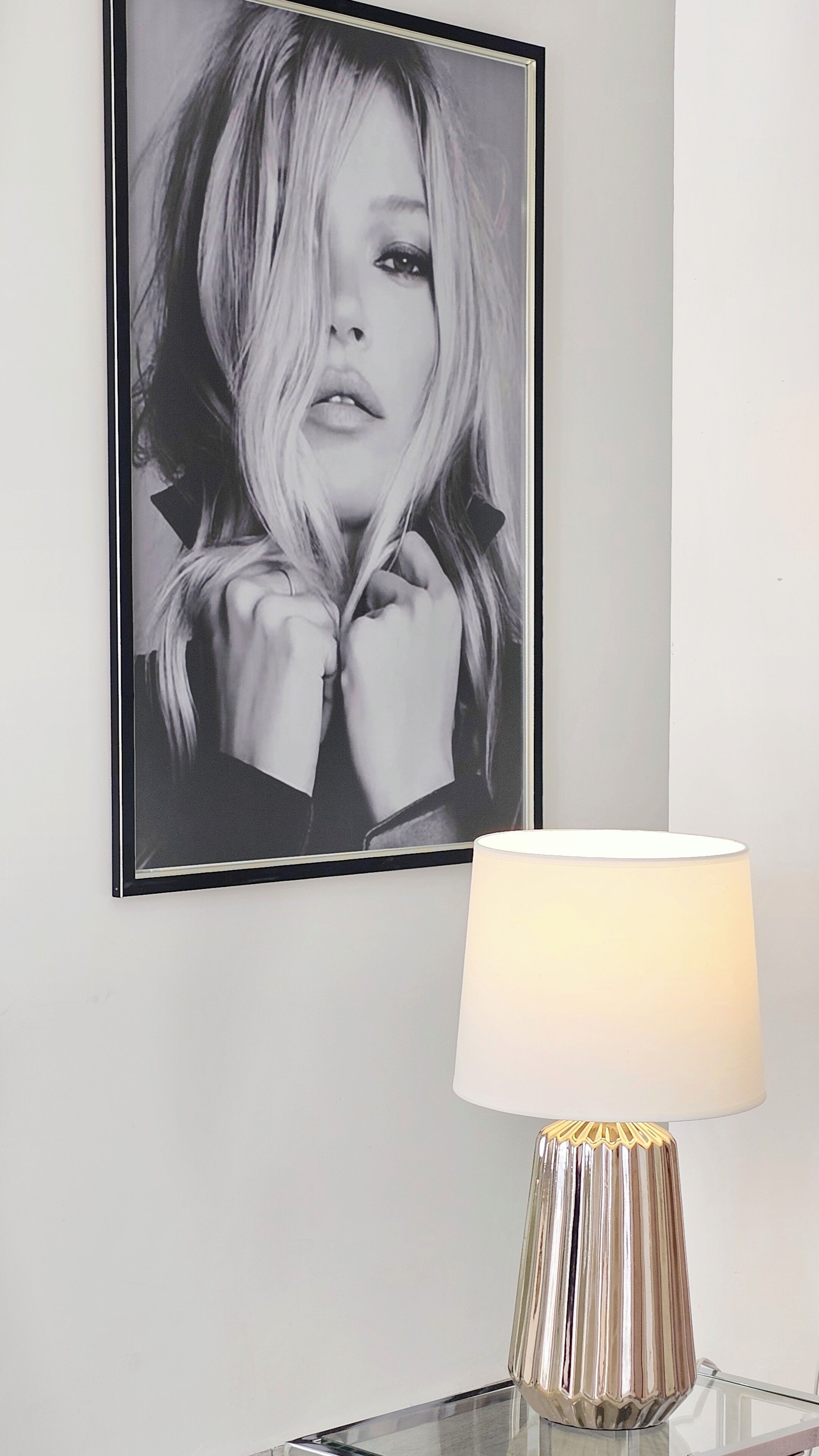 Une photographie en noir et blanc encadrée accrochée au mur, une lampe moderne avec un abat-jour blanc et un pied métallique, et une table en verre transparent.