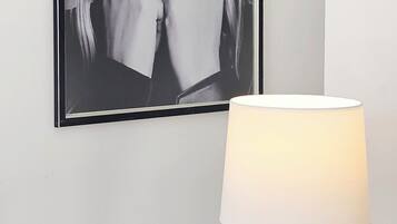 Une photographie en noir et blanc encadrée accrochée au mur, une lampe moderne avec un abat-jour blanc et un pied métallique, et une table en verre transparent.