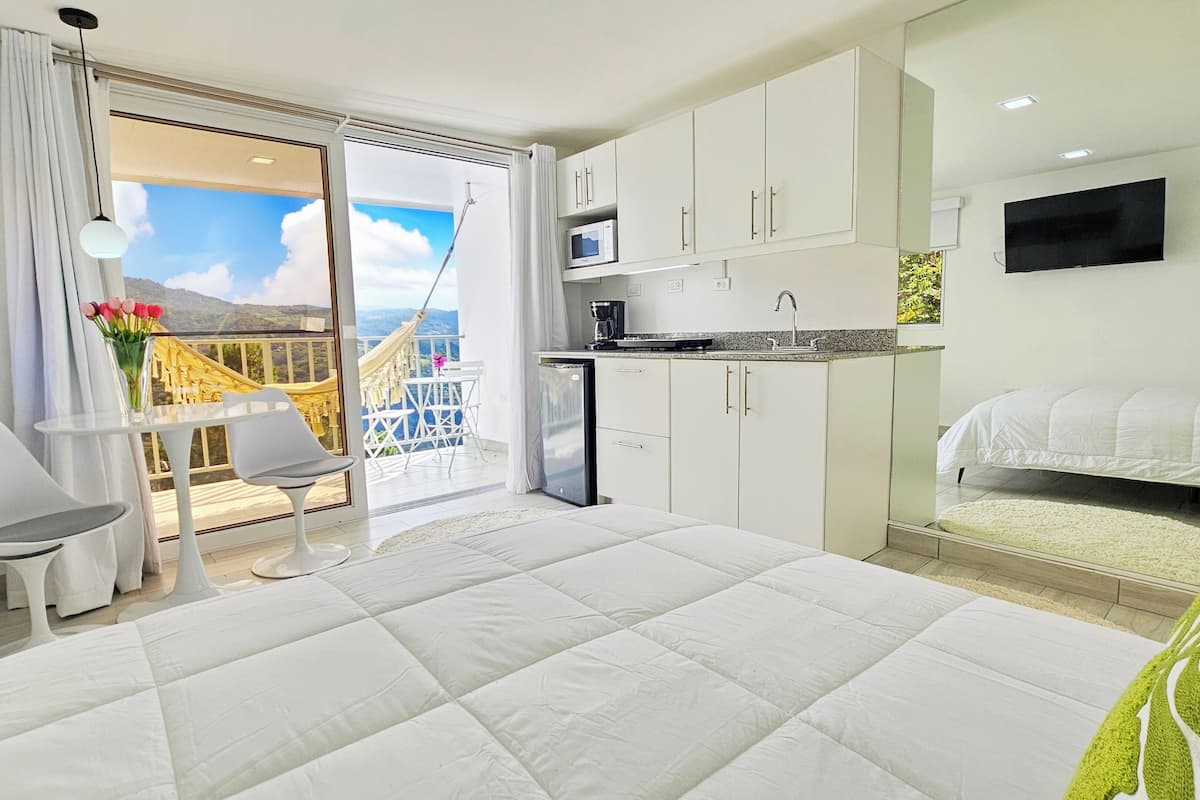 A modern hotel room with a balcony, a kitchenette, a bed, and a small table with chairs.