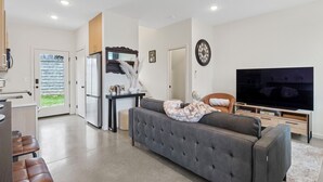 A modern living room with a grey sofa, a TV, and a kitchen area with a fridge and a dining table. - Back 2 The Future! - 4 Min. from Downtown Knox (Knoxville)