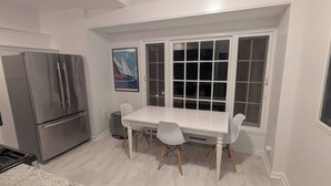 Ett kompakt kök med ett vitt bord och stolar, ett kylskåp i rostfritt stål och ett fönster med flera rutor.