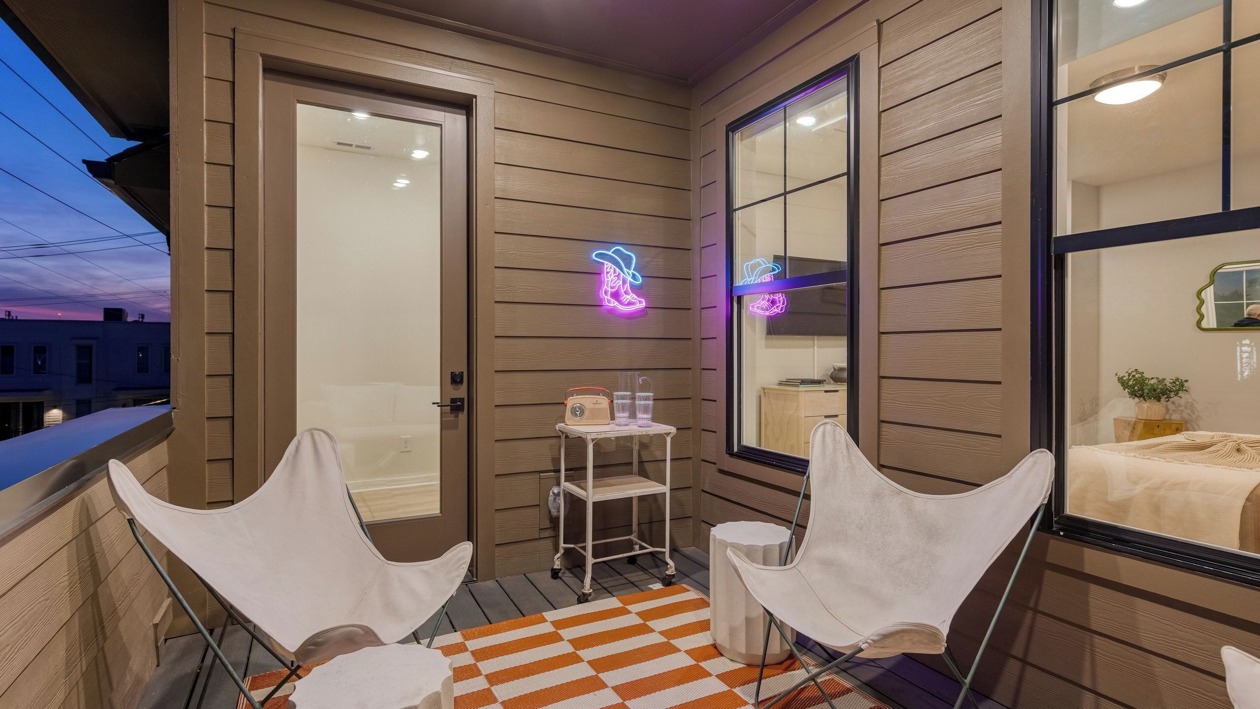 Un balcon avec deux chaises blanches, une petite table surmontée d’un néon et un tapis à carreaux.