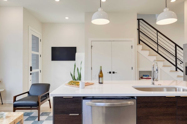 A modern kitchen with a central island, stainless steel dishwasher, and a built-in oven. There is a dining area with a table and chairs, and a staircase leading to an upper level. - Stunning New Build in East Nash Minutes to Broadway The Stage by Avantstay (Nashville)