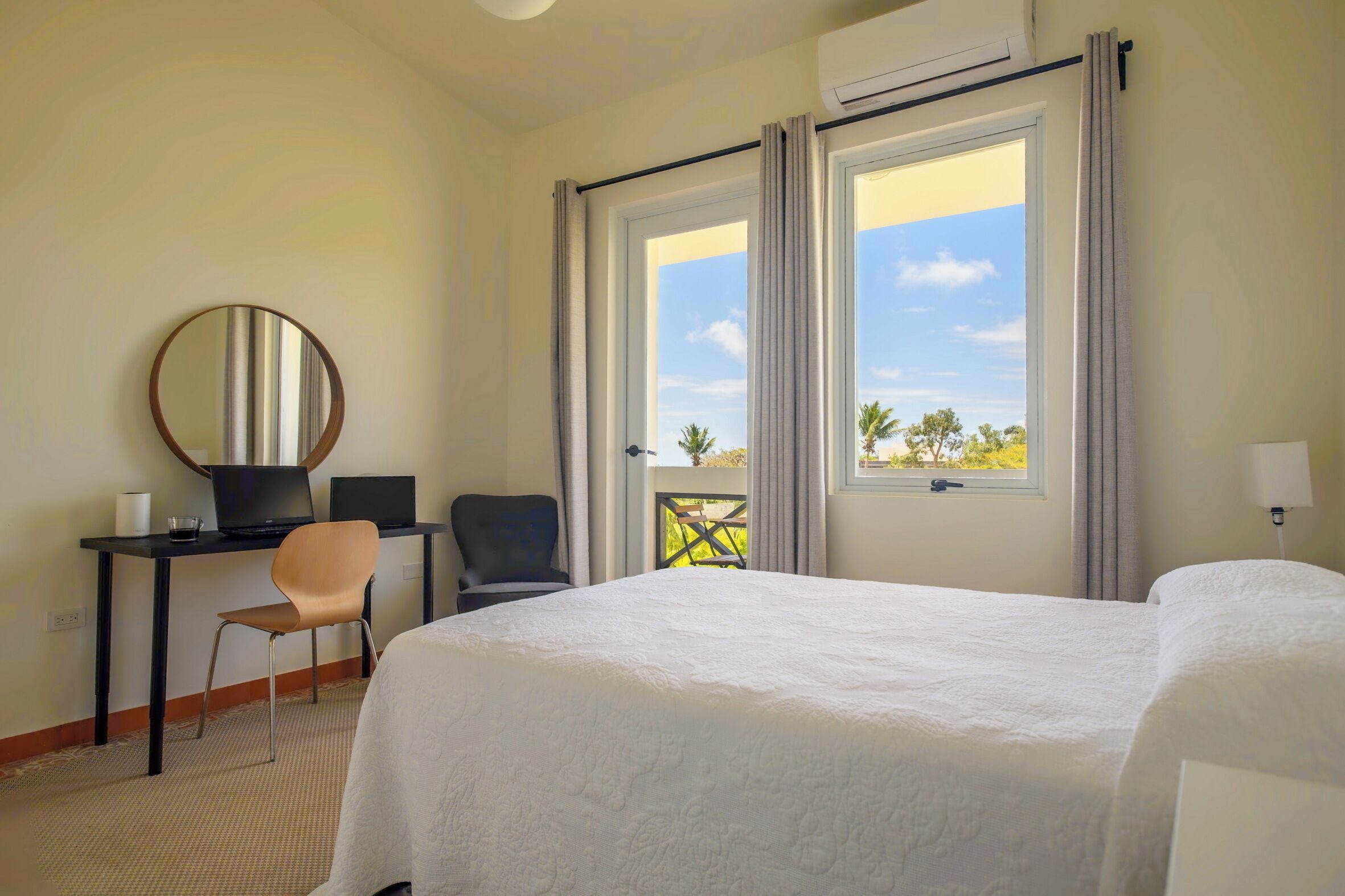 A hotel room with a bed, a desk with a laptop, a chair, a round mirror, and a balcony with a view of palm trees.