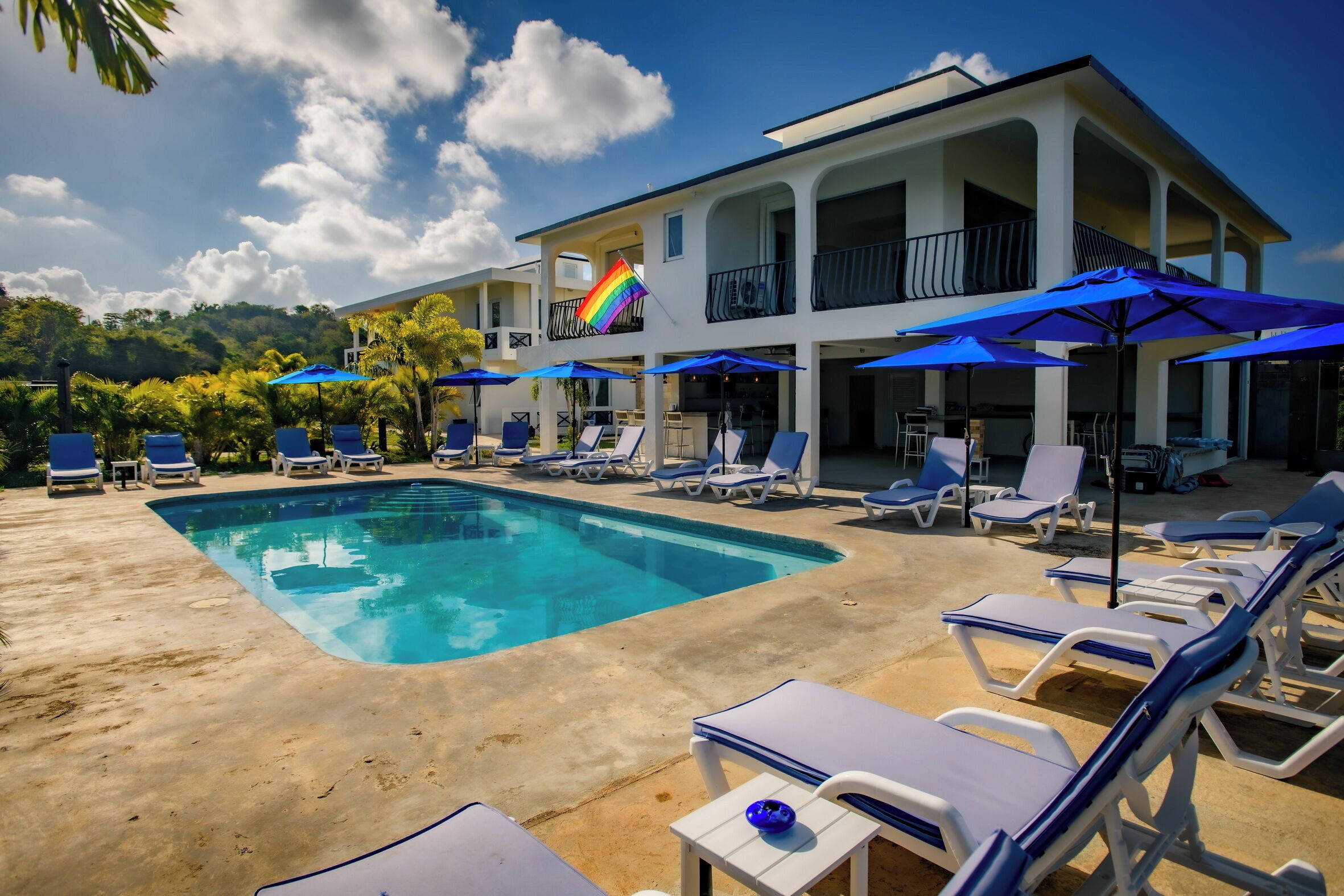 A pool area with lounge chairs and umbrellas, a two-story building with balconies, and a clear sky.