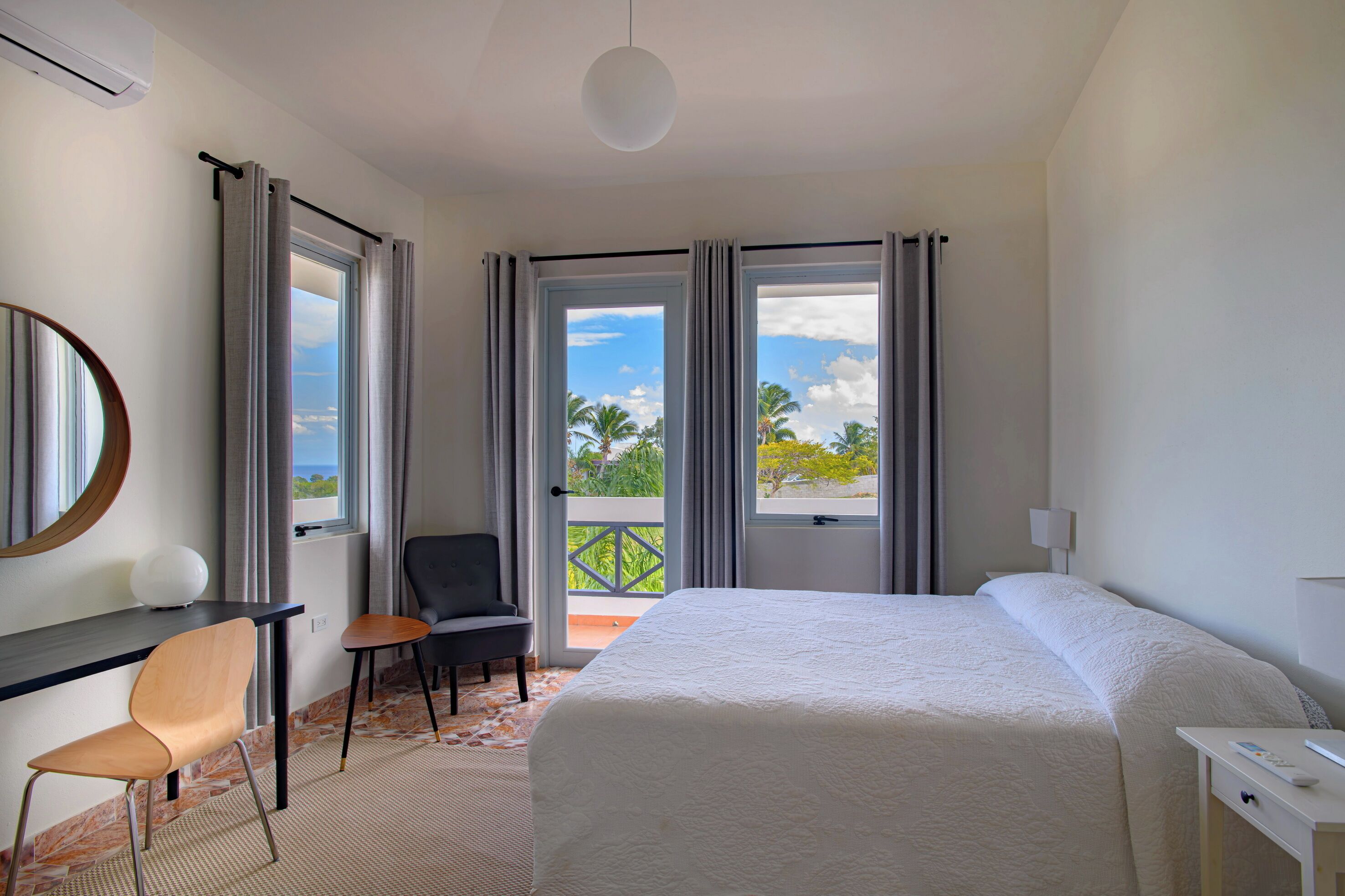 A hotel room with a bed, a desk, a chair, and a balcony with a view of palm trees and the sky.
