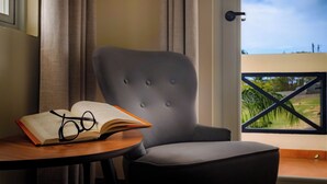 A room with a grey upholstered chair, a wooden table with an open book and glasses, and a window with a view of greenery. - Club Vieques - Adults Only Resort - LGBT Friendly (Vieques)