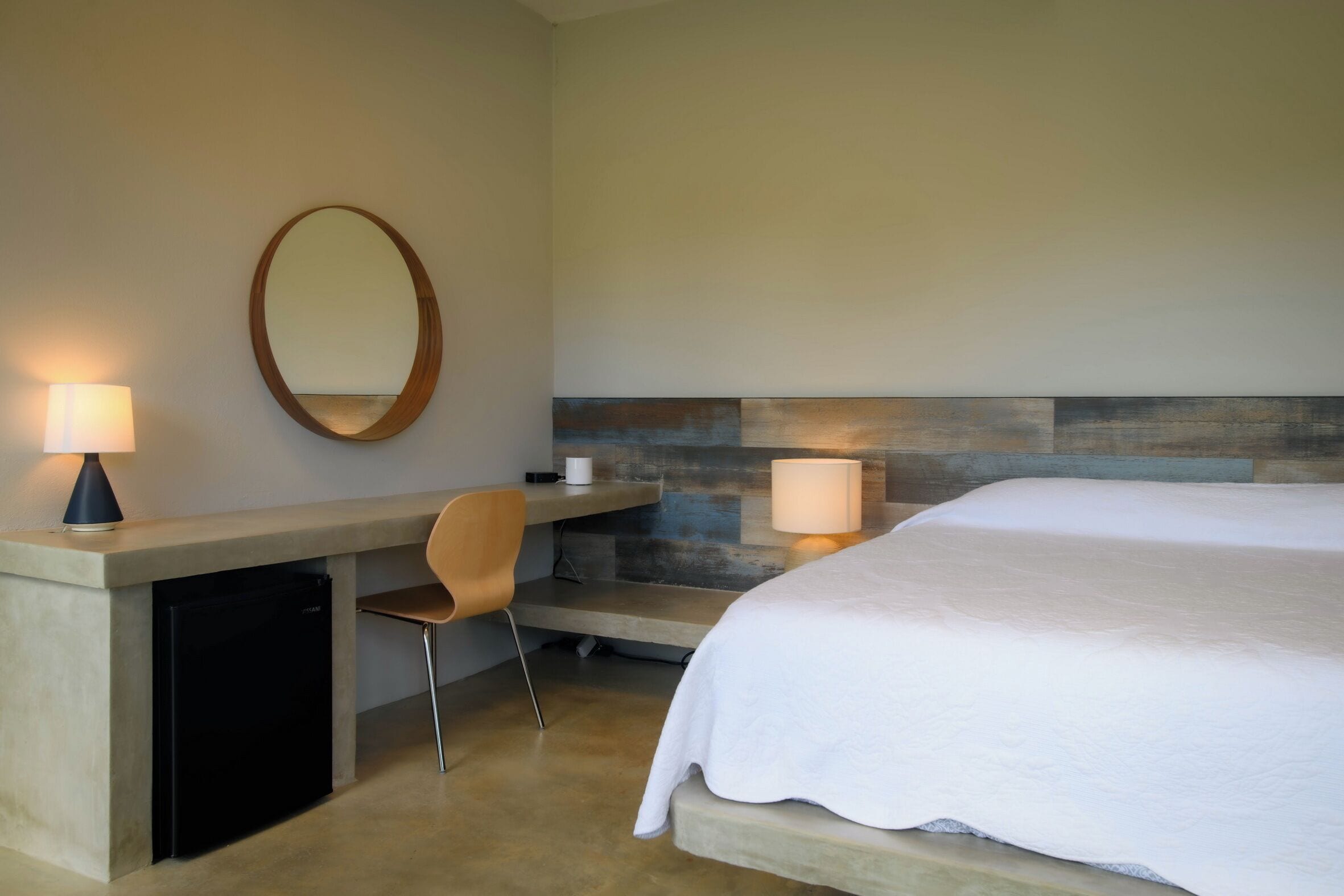 A hotel room with a bed, a desk with a chair, a small refrigerator, a round mirror, and two wall-mounted lamps.