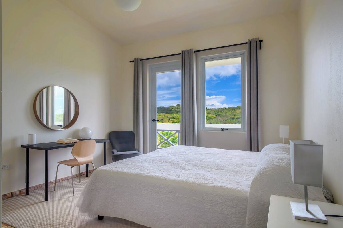A hotel room with a bed, a desk, a chair, a round mirror, and a window with curtains.