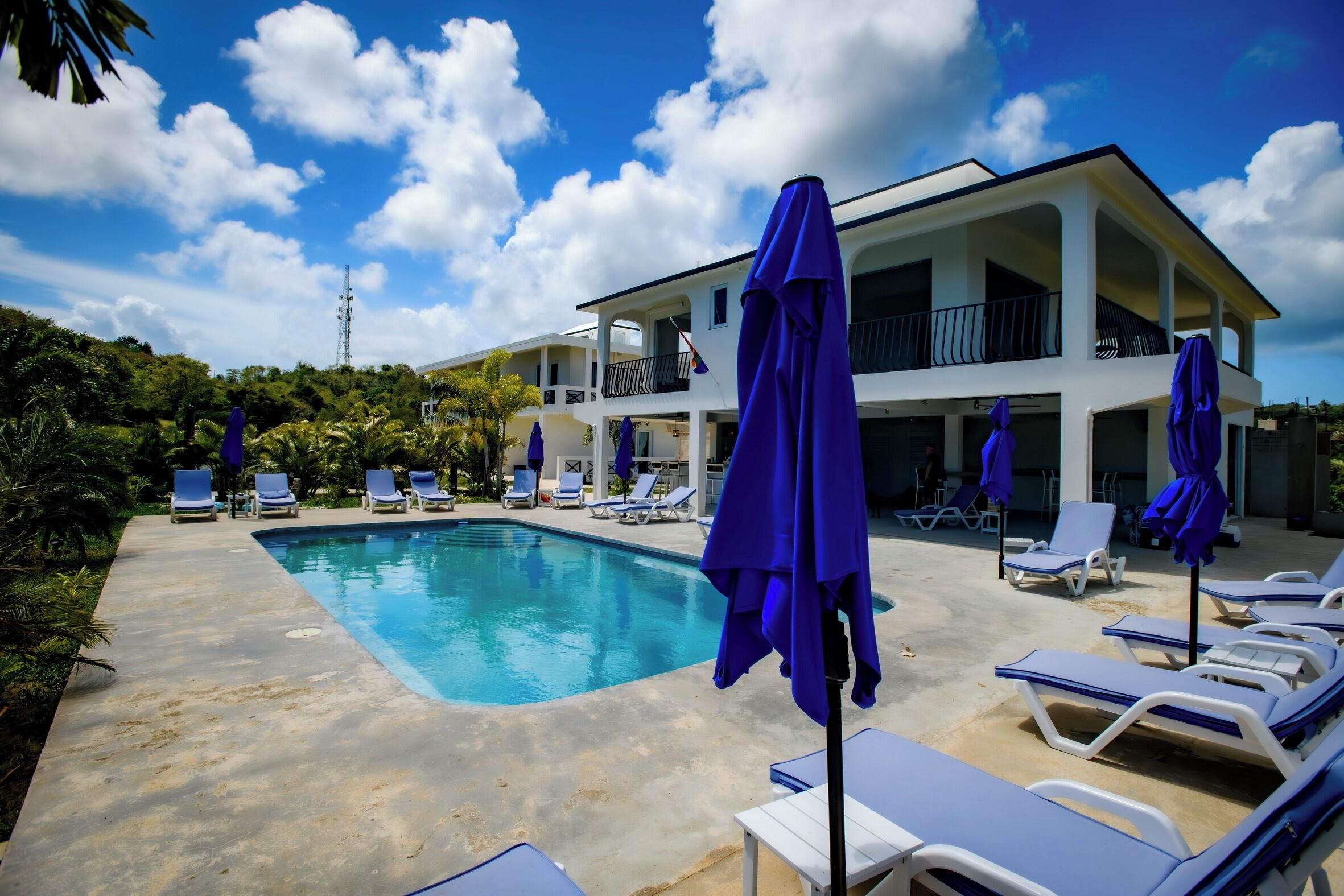 A pool area with lounge chairs and umbrellas, a two-story building with balconies, and a clear blue sky.