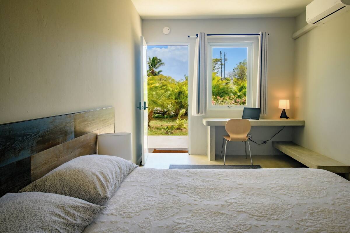 A bedroom with a large bed, a desk with a chair, and a view of greenery outside.