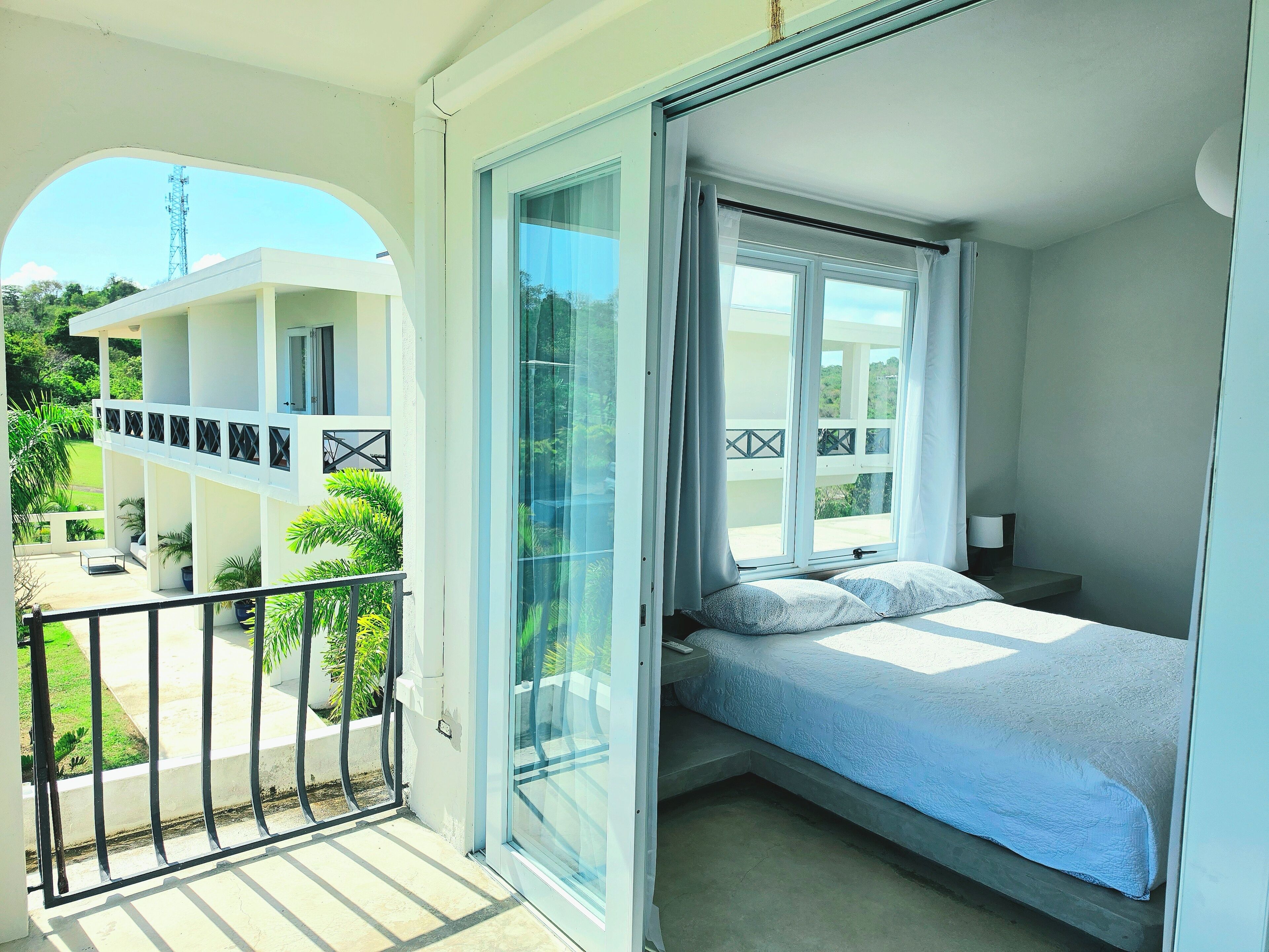 A balcony with a view of a building and greenery, a bed with pillows, and a nightstand with a lamp.