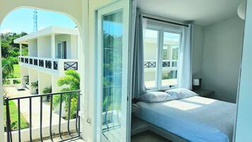 A balcony with a view of a building and greenery, a bed with pillows, and a nightstand with a lamp.