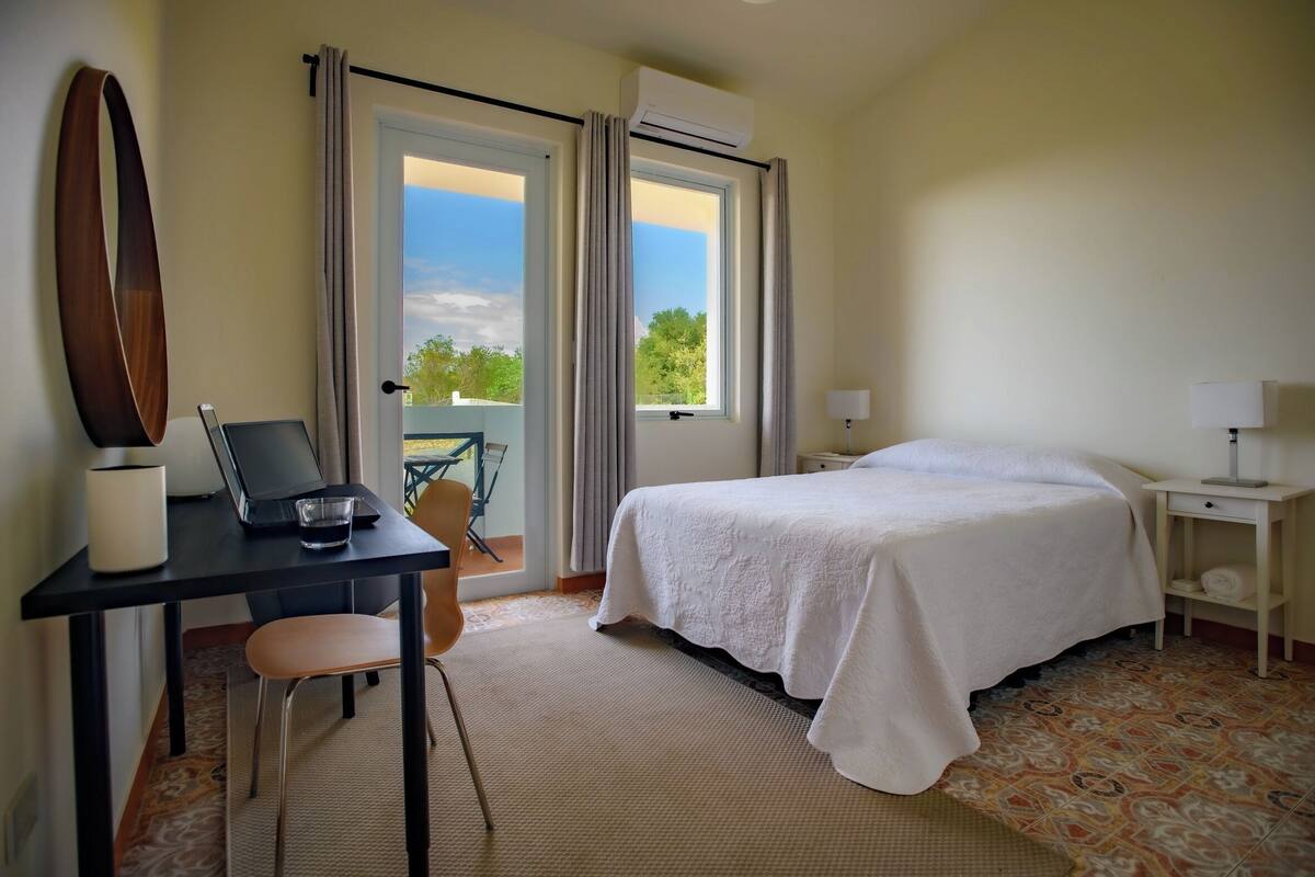 A hotel room with a bed, a desk with a laptop, a chair, a lamp, and a view of the outdoors.