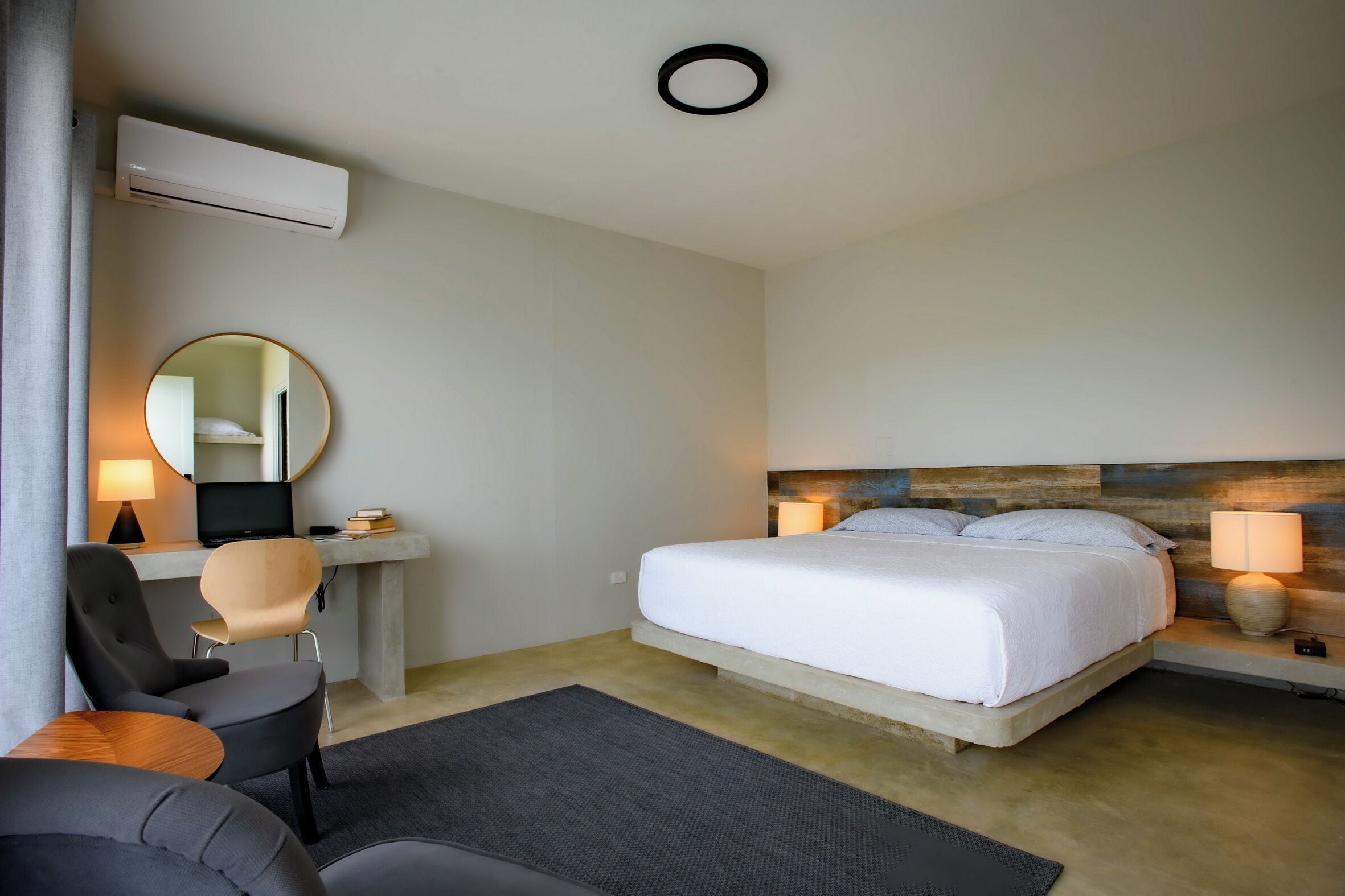 A modern hotel room with a bed, a desk with a chair, a round mirror, and a bedside table with a lamp.