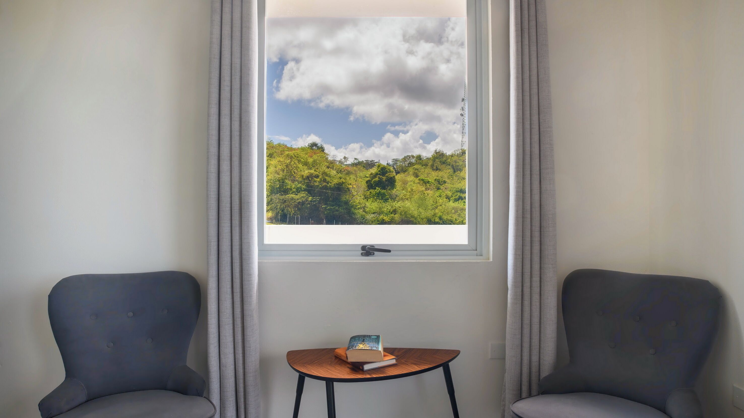 Ein Zimmer mit zwei grauen Sesseln, einem kleinen Holztisch mit einem Buch und einem Fenster mit Blick auf einen grünen Wald und blauen Himmel.
