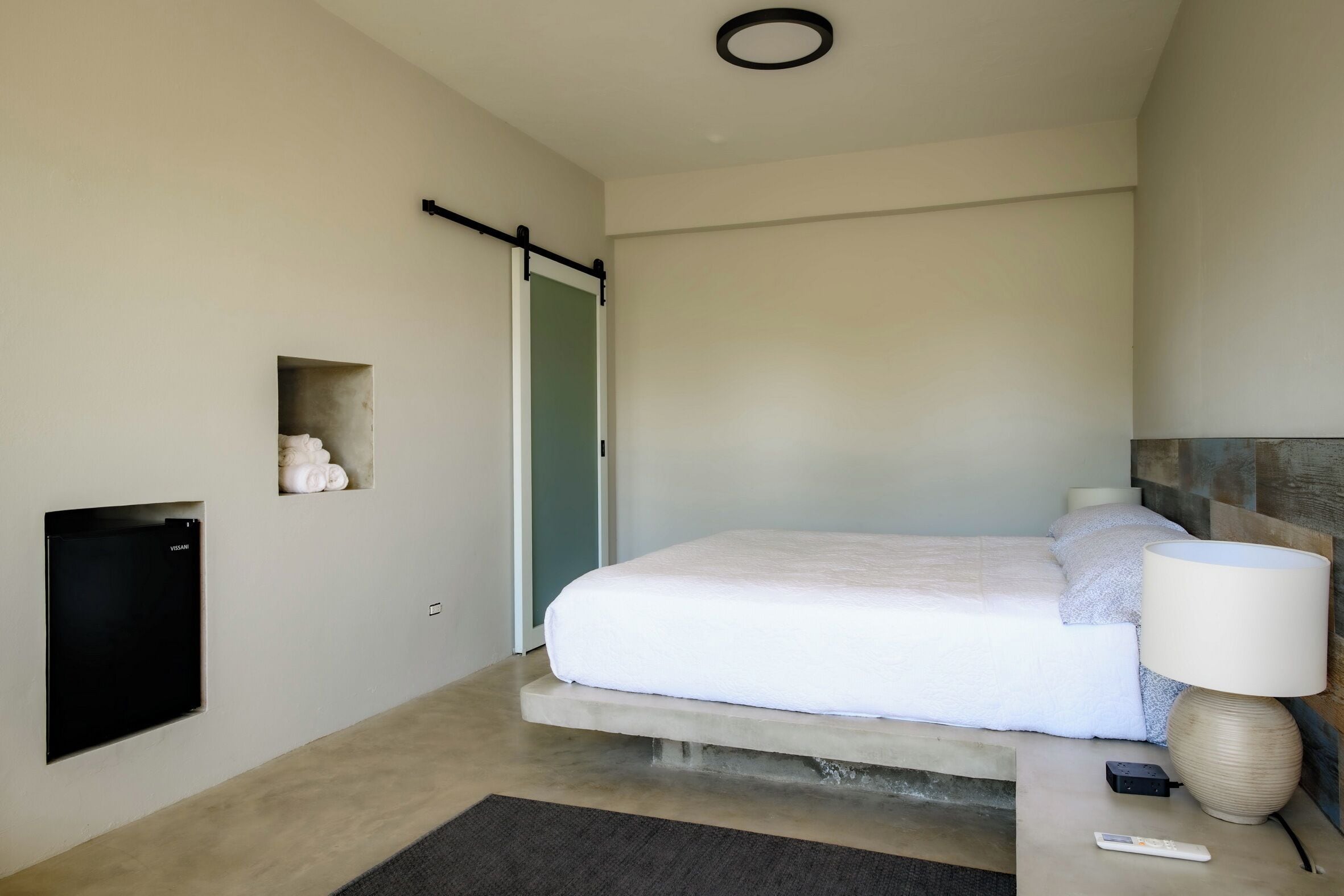 Habitació d'hotel moderna amb un llit, una làmpada de nit, una televisió i una petita estant amb objectes decoratius.
