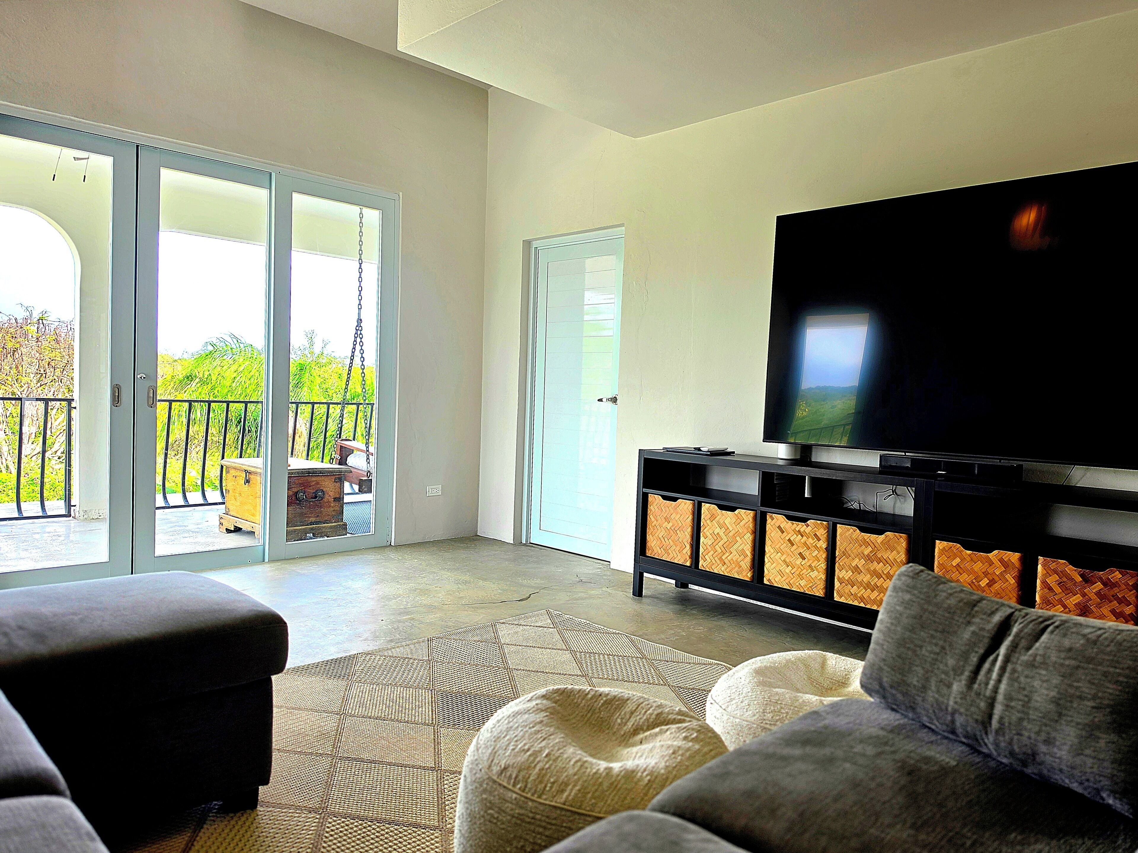 A living room with a flat-screen TV, a sofa, ottomans, and a balcony with a view.