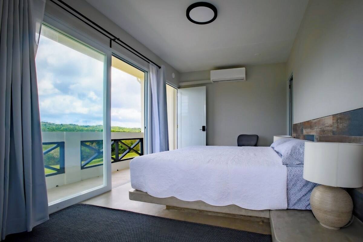 A bedroom with a large bed, a chair, a bedside table, a lamp, and a view of a balcony with a railing.