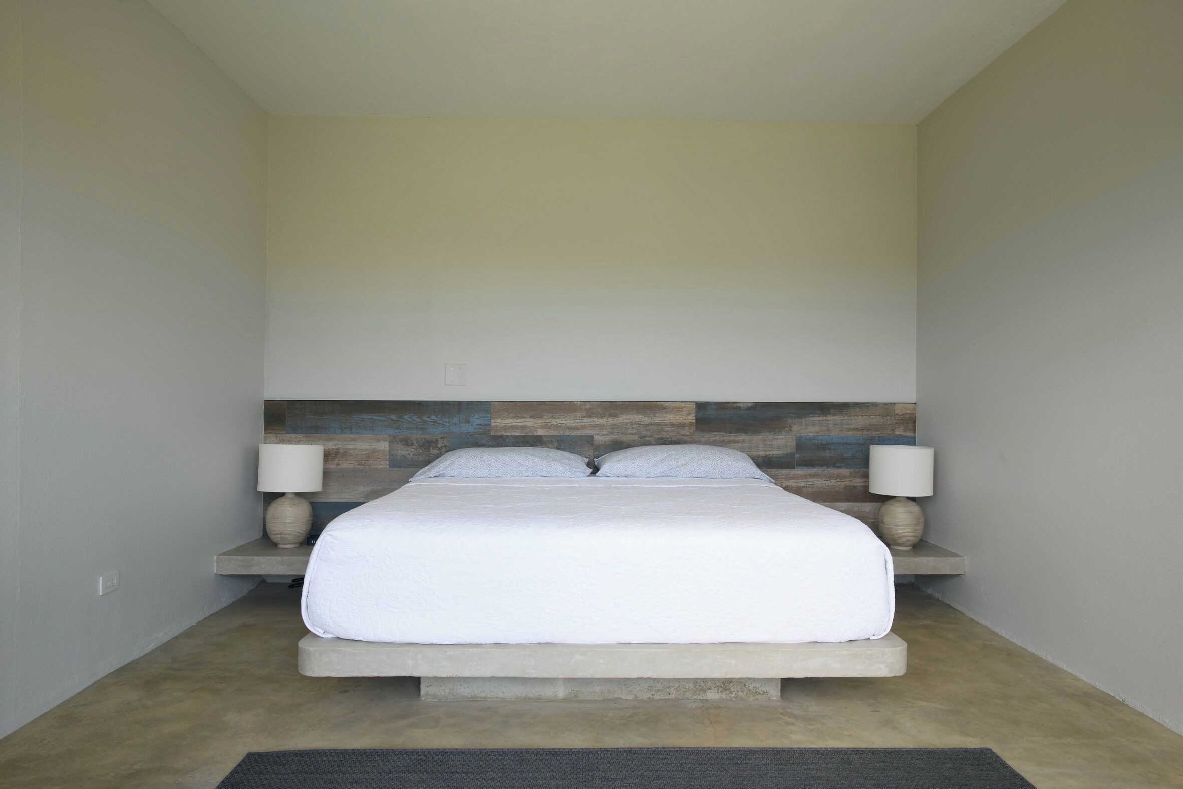 Un dormitorio con una cama centrada sobre una plataforma, flanqueada por dos mesitas de noche con lámparas, frente a una pared con diseño de paneles de madera.