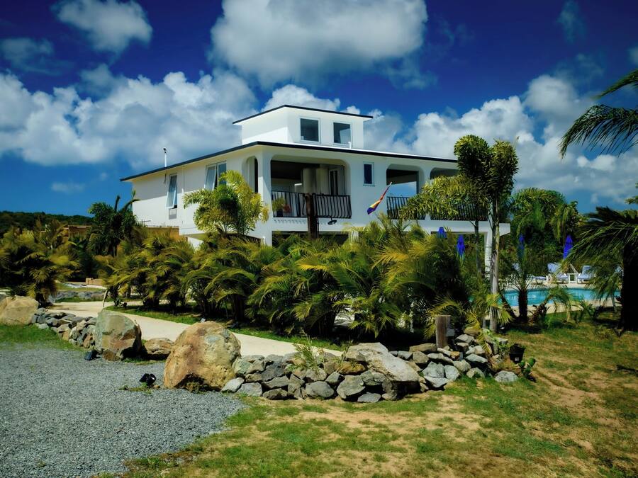 A white two-story house with a balcony, surrounded by palm trees and a swimming pool.