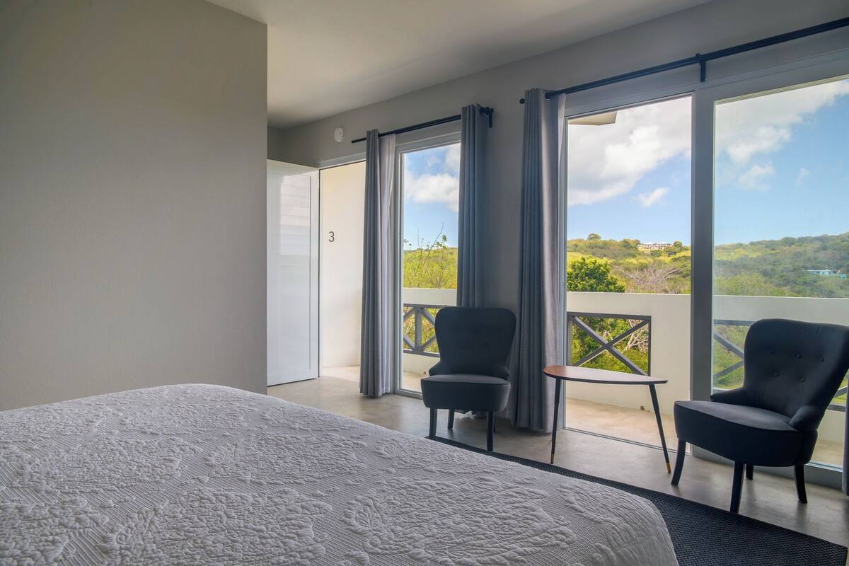 A bedroom with a large bed, two chairs, a small table, and a balcony with a view of greenery.