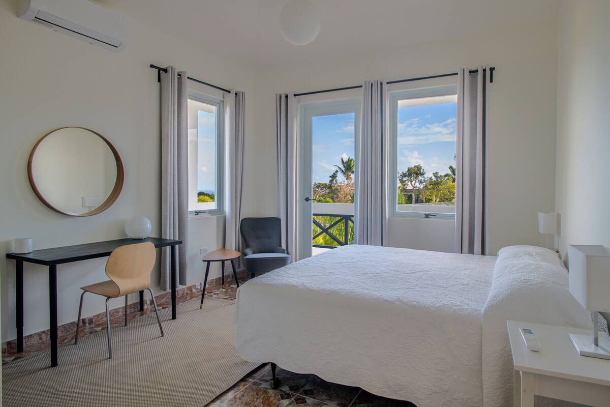 A hotel room with a large bed, a desk with a chair, a round mirror, and a view of the outdoors through the windows.