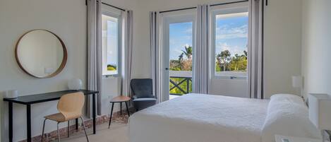 A hotel room with a large bed, a desk with a chair, a round mirror, and a view of the outdoors through the windows.