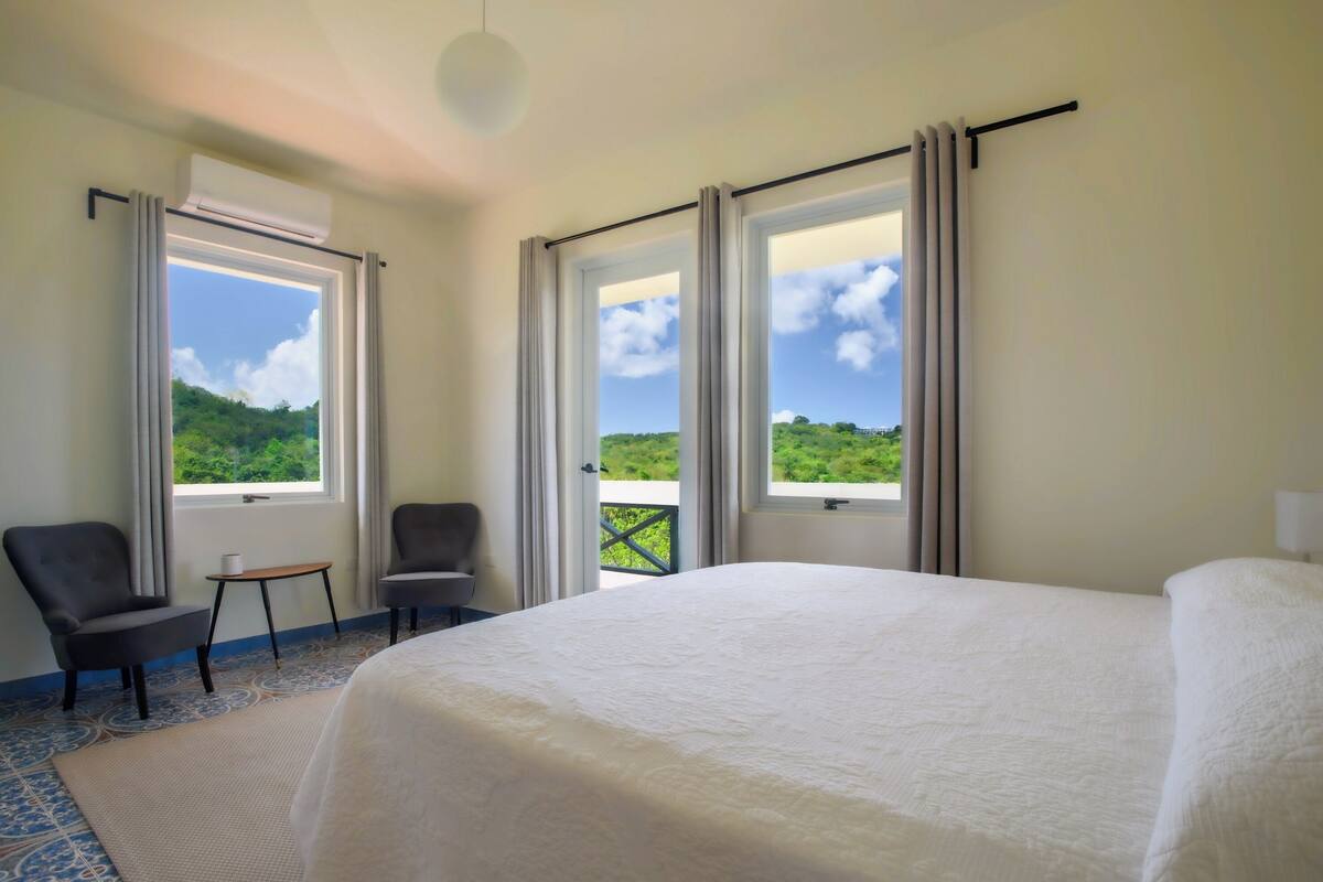 A hotel room with a large bed, two chairs, a small table, and a view of a green landscape through the windows.