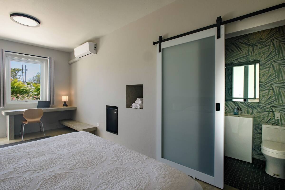 A bedroom with a bed, a desk, a chair, a sliding door to a bathroom, and a window with a view of greenery.