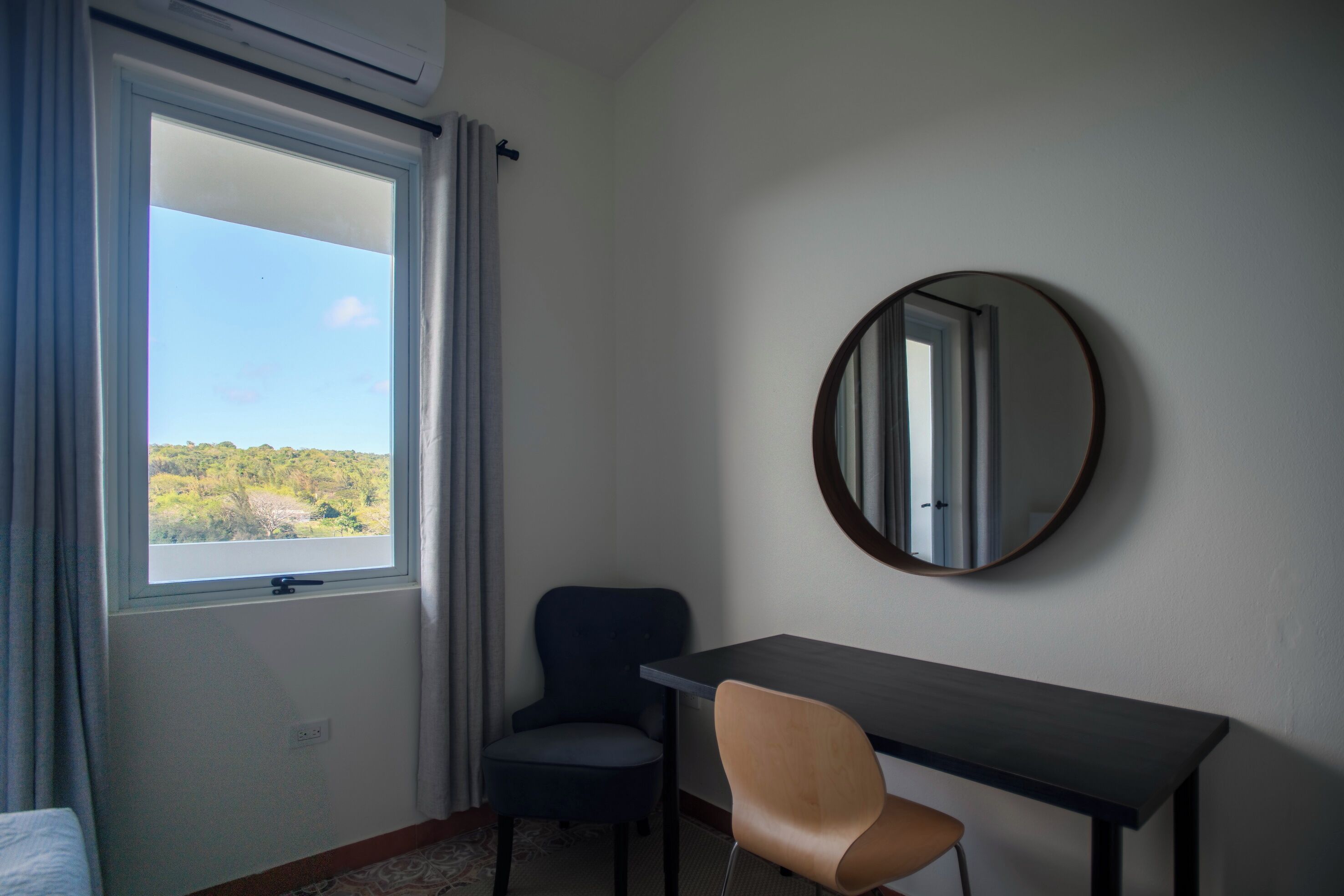 A room with a bed, a desk, a chair, a round mirror, and a window with curtains.