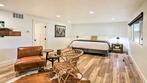 Un dormitorio con una cama, un sillón de cuero marrón, una silla de mimbre, una mesita redonda y un tapete de cuero de vaca.
